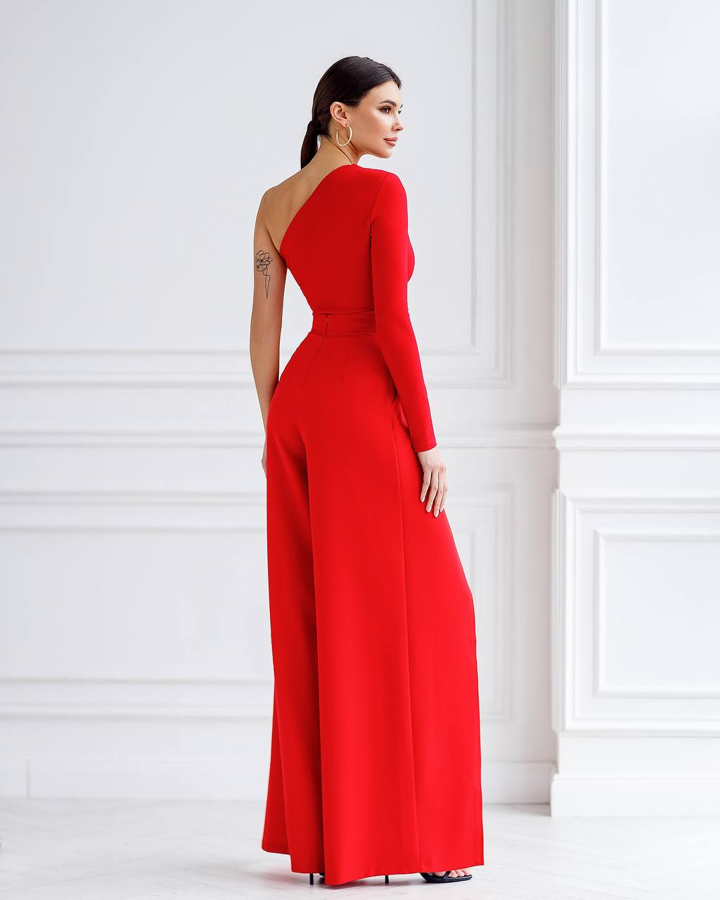 Woman wearing a red one-shoulder jumpsuit against a white wall.