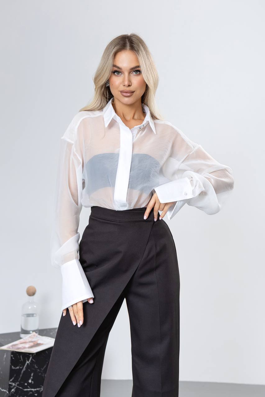 Woman wearing a white sheer blouse with a blue pattern and black pants against a plain background