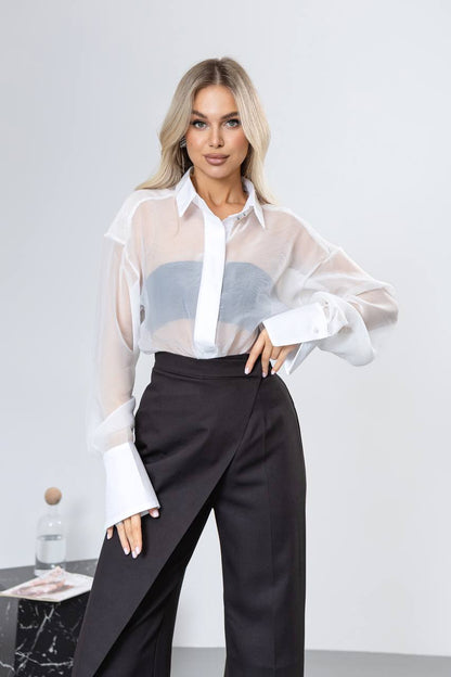 Woman wearing a white sheer blouse with a blue pattern and black pants against a plain background