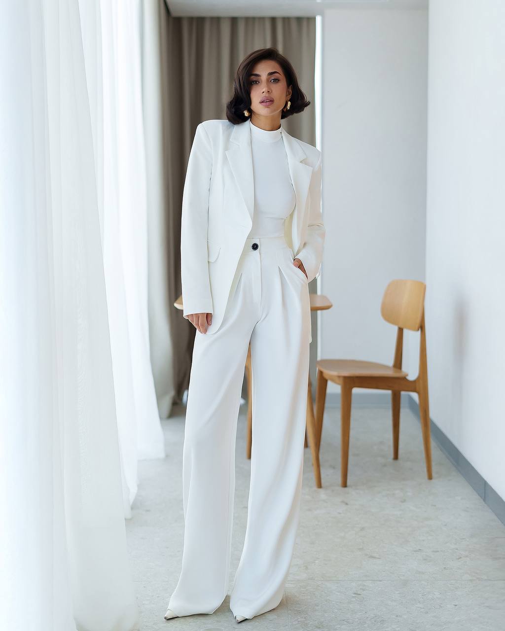 Woman in a white suit standing in a minimalistic room with light-colored walls and a wooden chair.