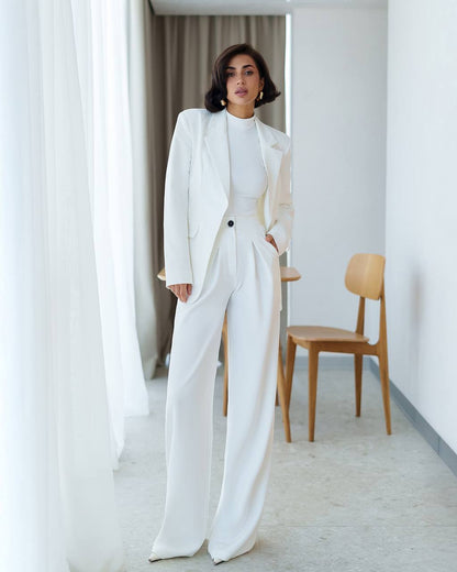Woman in a white suit standing in a minimalistic room with light-colored walls and a wooden chair.