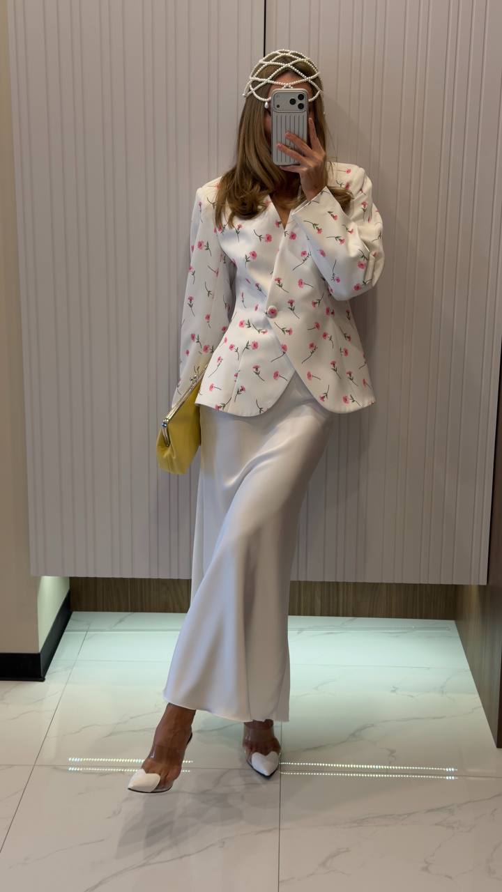 Person wearing a white floral blouse and white pants with a yellow handbag, standing in a hallway.