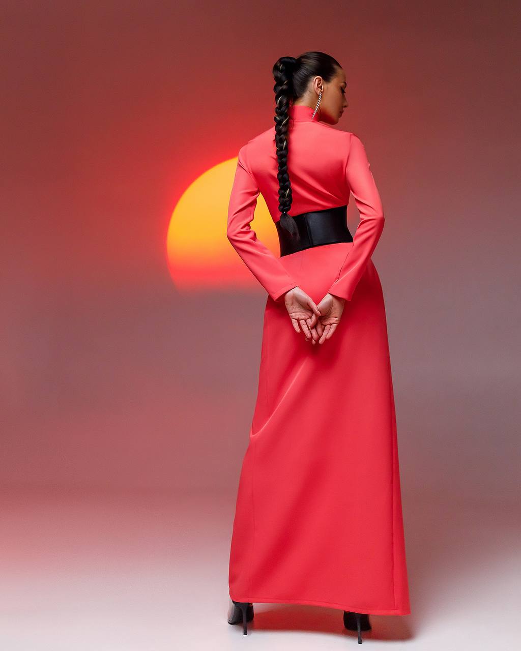 Woman in a long red dress with a black belt standing against a gradient background with a sun-like shape.
