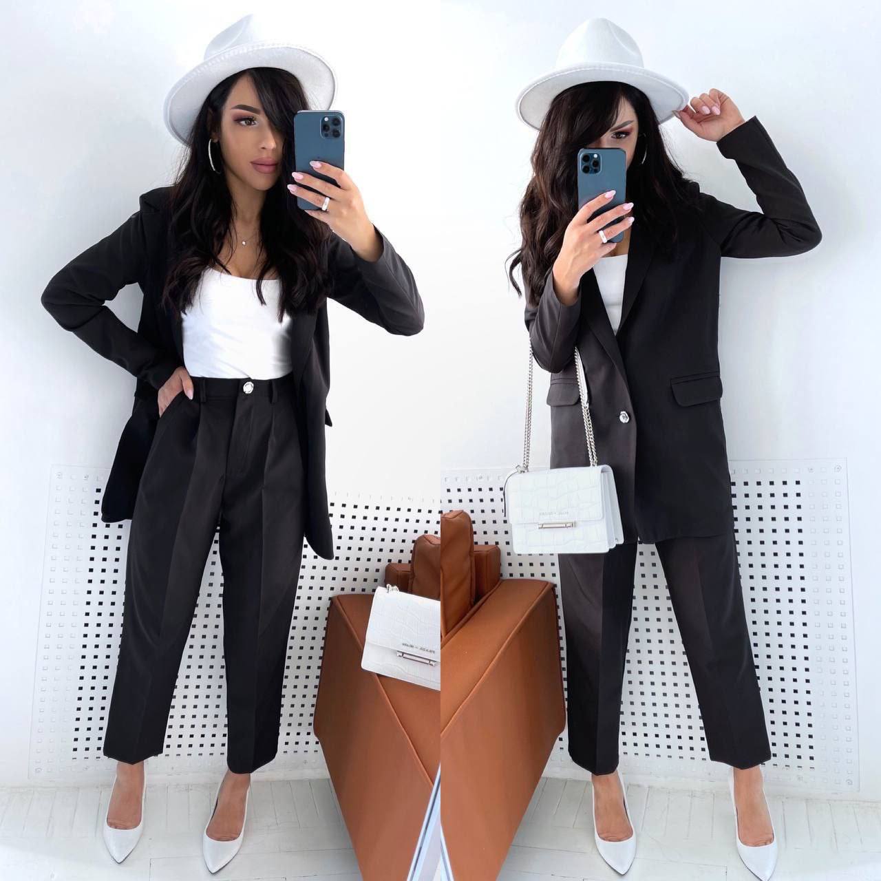 Woman in a black suit and white hat taking a mirror selfie against a white wall.