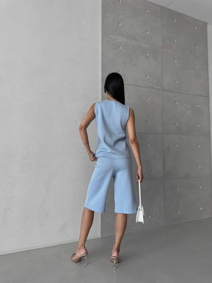 Woman in a light blue sleeveless top and shorts standing against a gray concrete wall.