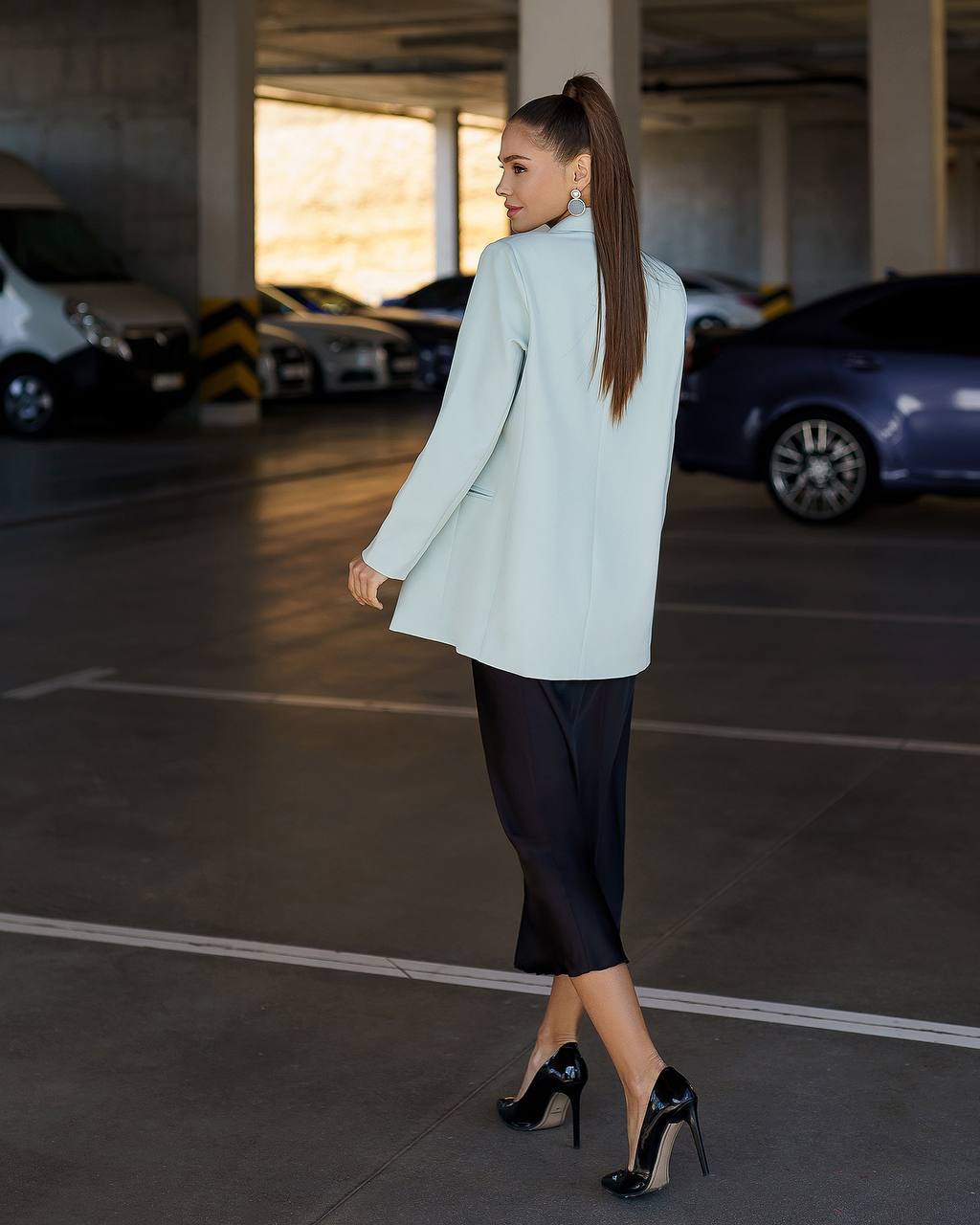 A woman wearing a light mint green blazer, black midi skirt, and black patent stiletto heels walks through a modern parking garage.