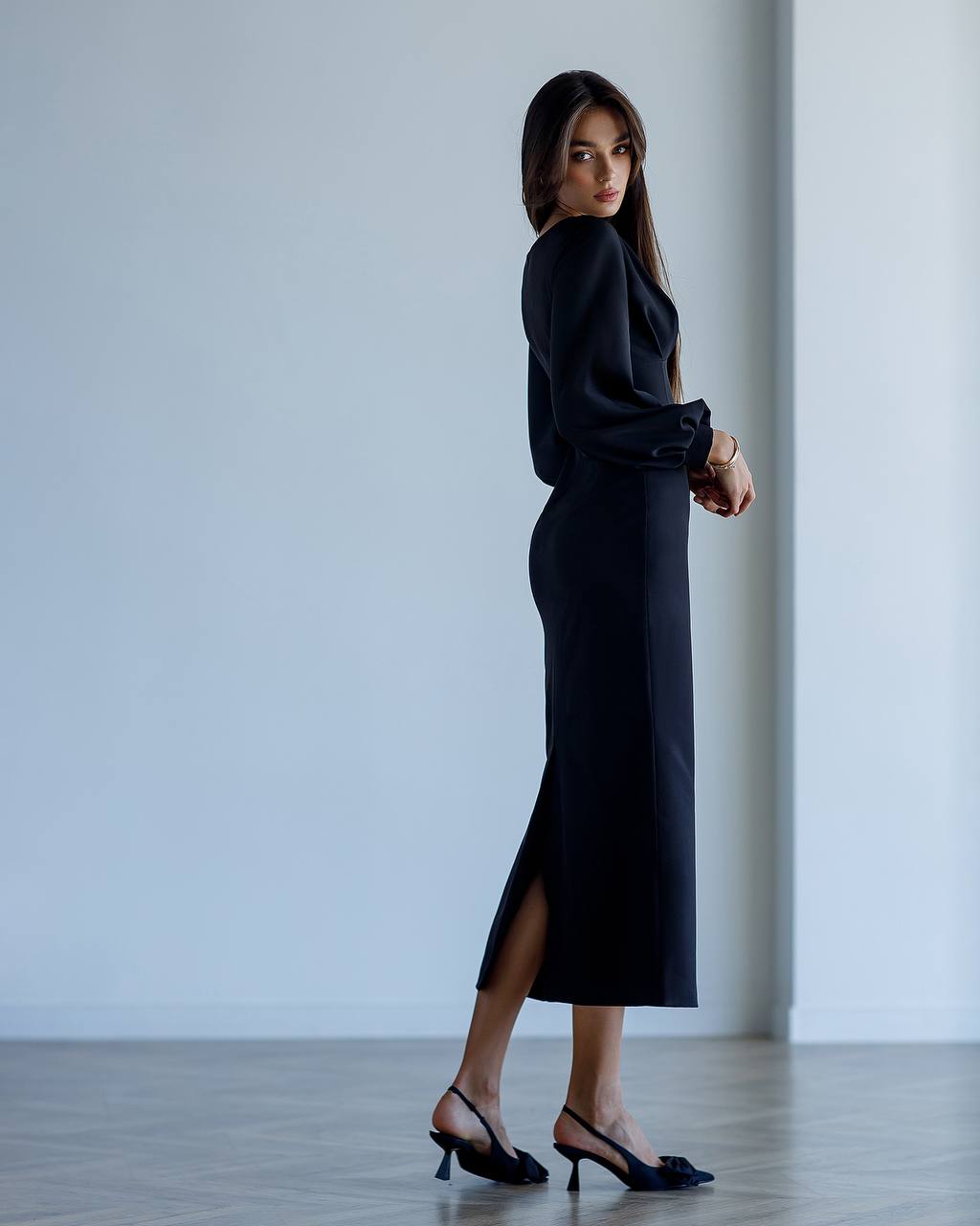 Woman wearing a black dress with a slit, standing against a plain white wall.