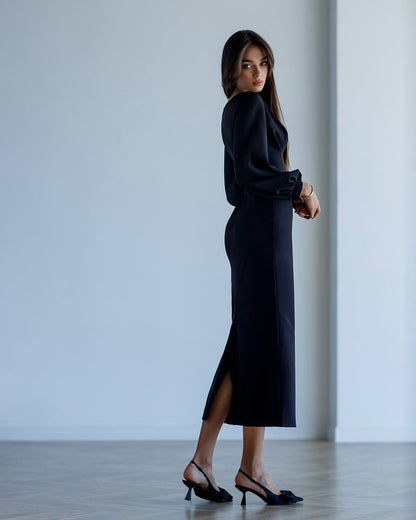 Woman wearing a black dress with a slit, standing against a plain white wall.