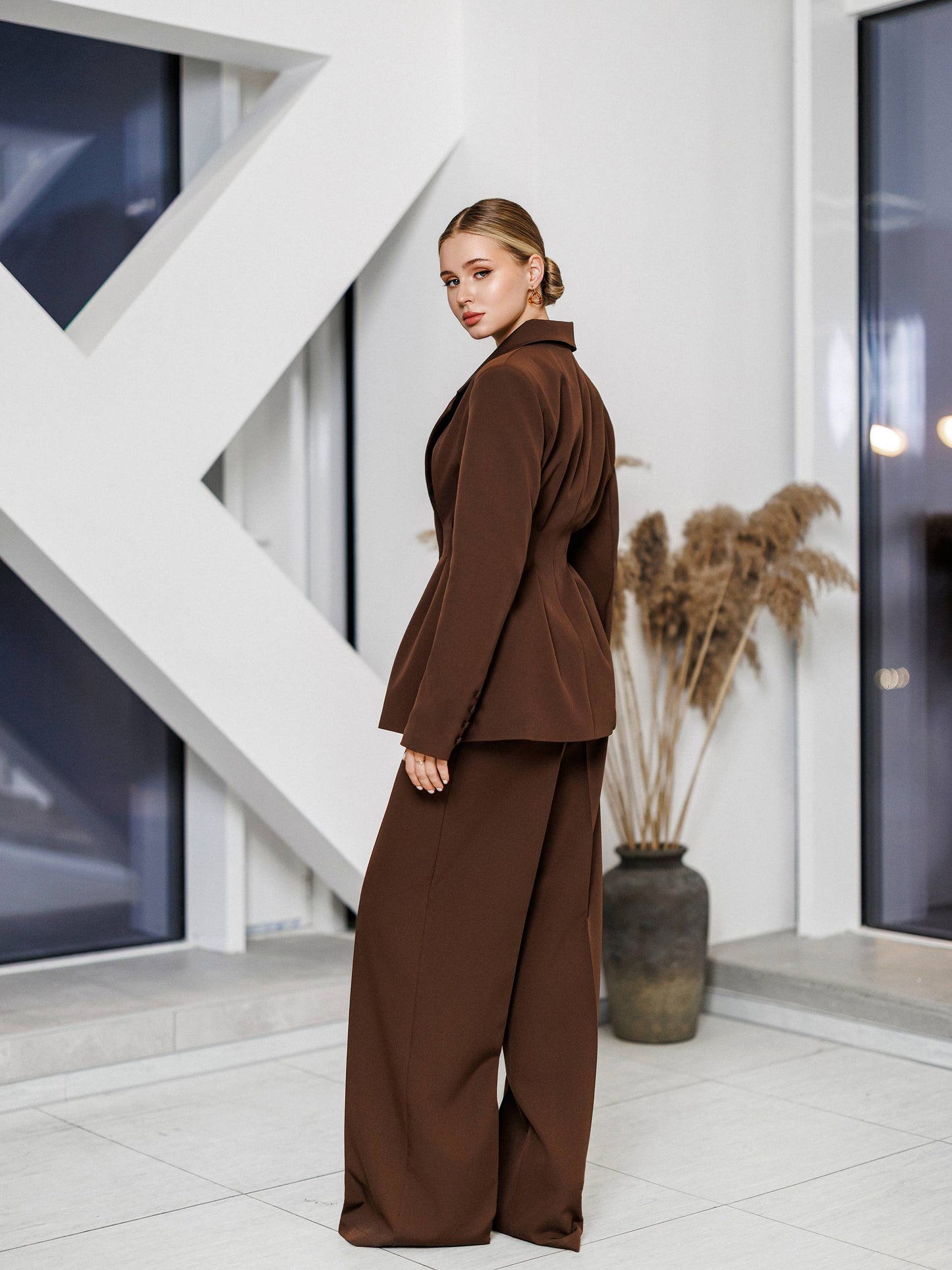 Woman in a brown suit standing in a modern indoor setting with white walls and geometric design.