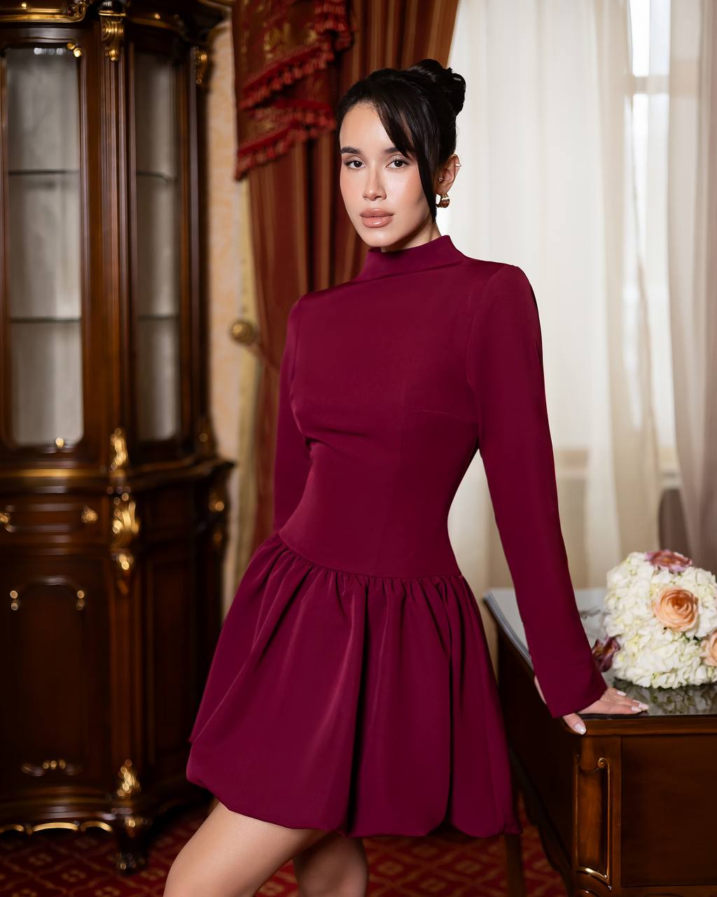 A woman wearing a long-sleeve, high-neck, fitted mini dress in deep burgundy, posing in an elegant room with a dark wood cabinet and a side table with flowers.