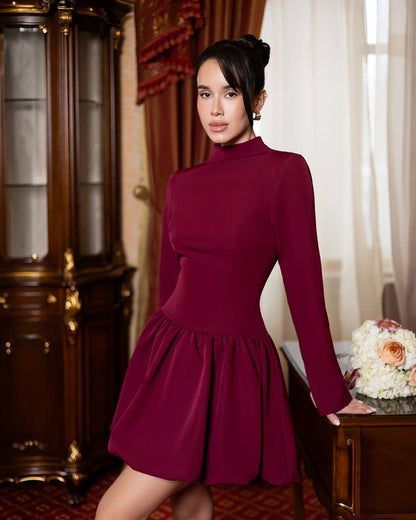 A woman wearing a long-sleeve, high-neck, fitted mini dress in deep burgundy, posing in an elegant room with a dark wood cabinet and a side table with flowers.