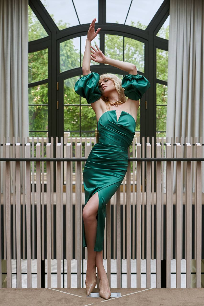 Woman in a green dress posing in front of a large window with a wooden slat screen.