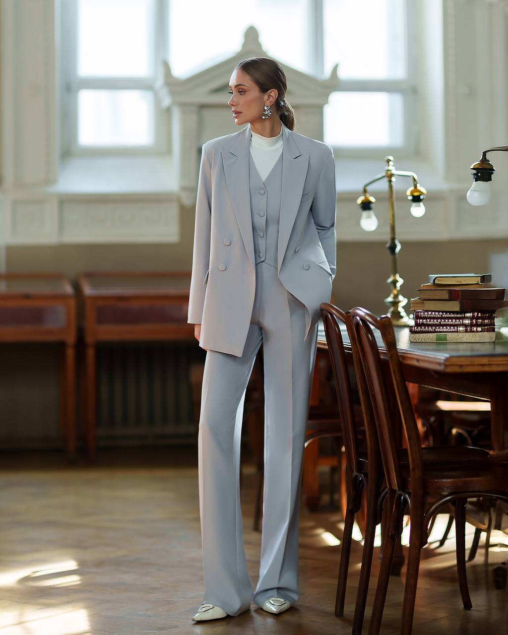 Woman in a gray suit standing in a room with wooden furniture and large windows.