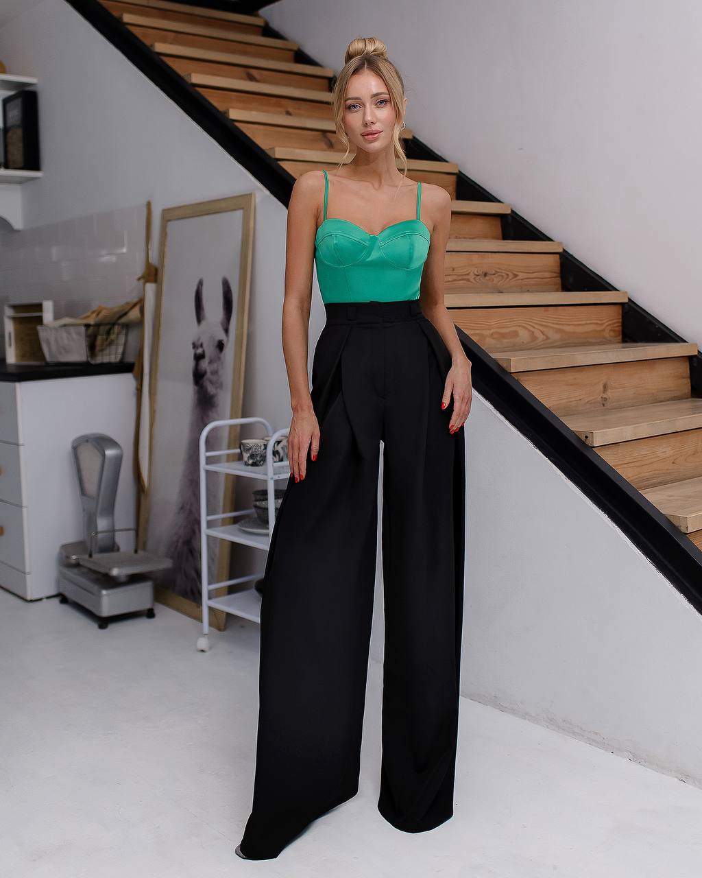 Woman in green top and black pants standing in a room with wooden stairs and decor.