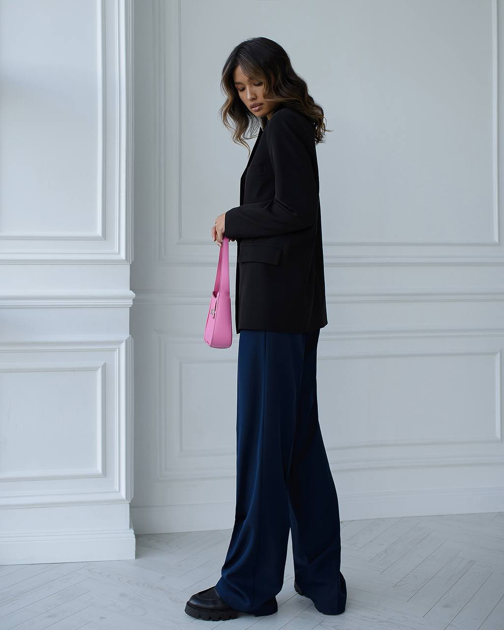 Person wearing a black blazer and blue pants holding a pink bag against a white paneled wall.