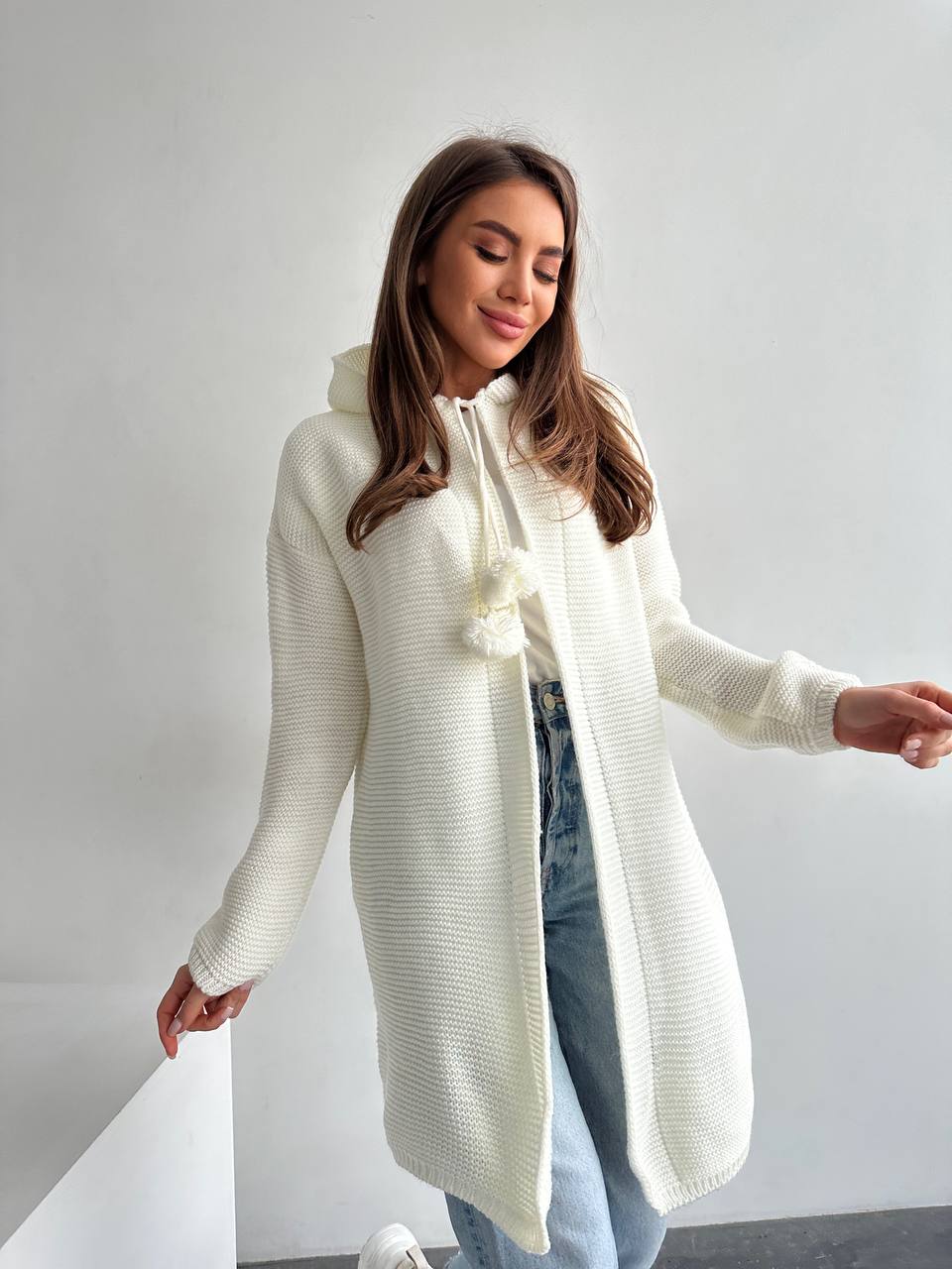 Woman wearing a long cream cardigan and light blue jeans against a plain background