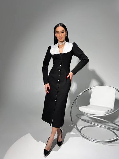 Woman in a black dress with white collar standing next to a modern chair on a gray background