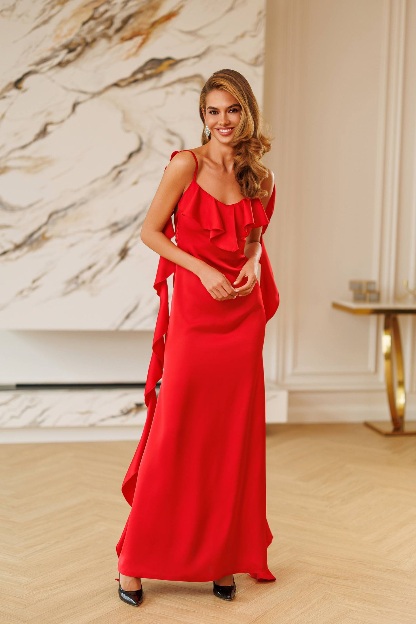 A woman modeling a vibrant red one-shoulder evening gown with a ruffled neckline and a flowing skirt, standing in a modern interior with a marble wall and light wood floors.