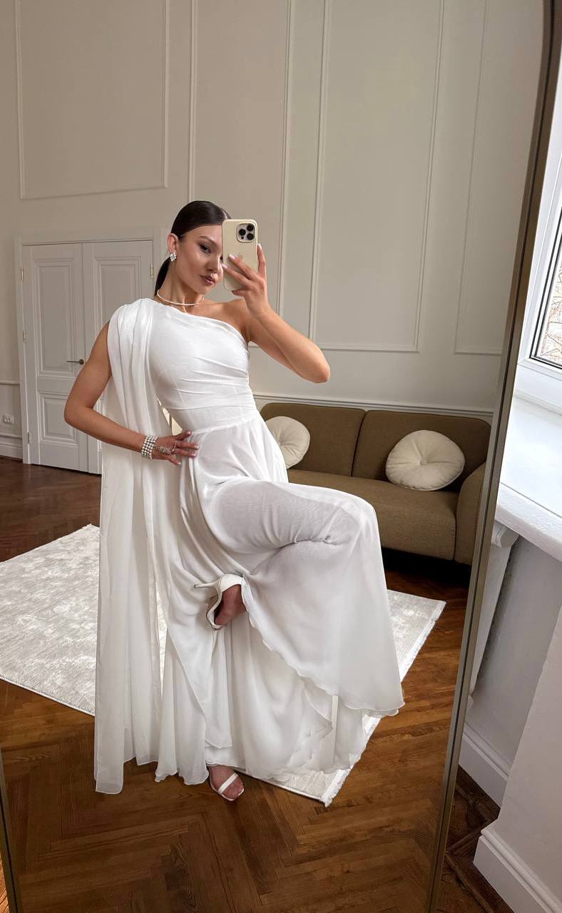 Woman in a white dress taking a mirror selfie in a room with a couch and decorative pillows.