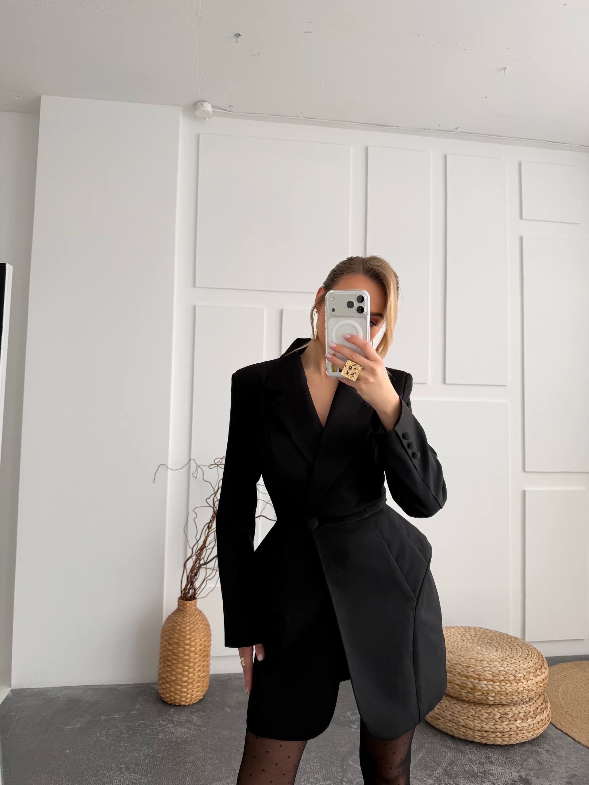 A woman takes a mirror selfie wearing a black asymmetrical blazer dress with a wrap-style front, paired with sheer black tights. The setting is a minimalist room with white paneled walls, a gray floor, and woven decorative elements.