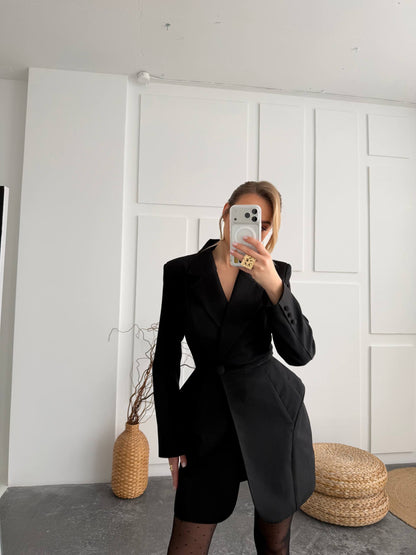 A woman takes a mirror selfie wearing a black asymmetrical blazer dress with a wrap-style front, paired with sheer black tights. The setting is a minimalist room with white paneled walls, a gray floor, and woven decorative elements.