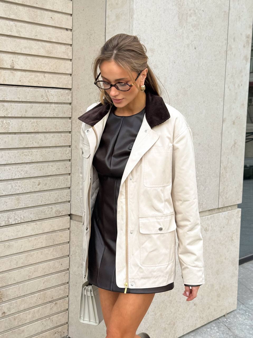 Woman wearing a white coat with a black collar against a light-colored wall.