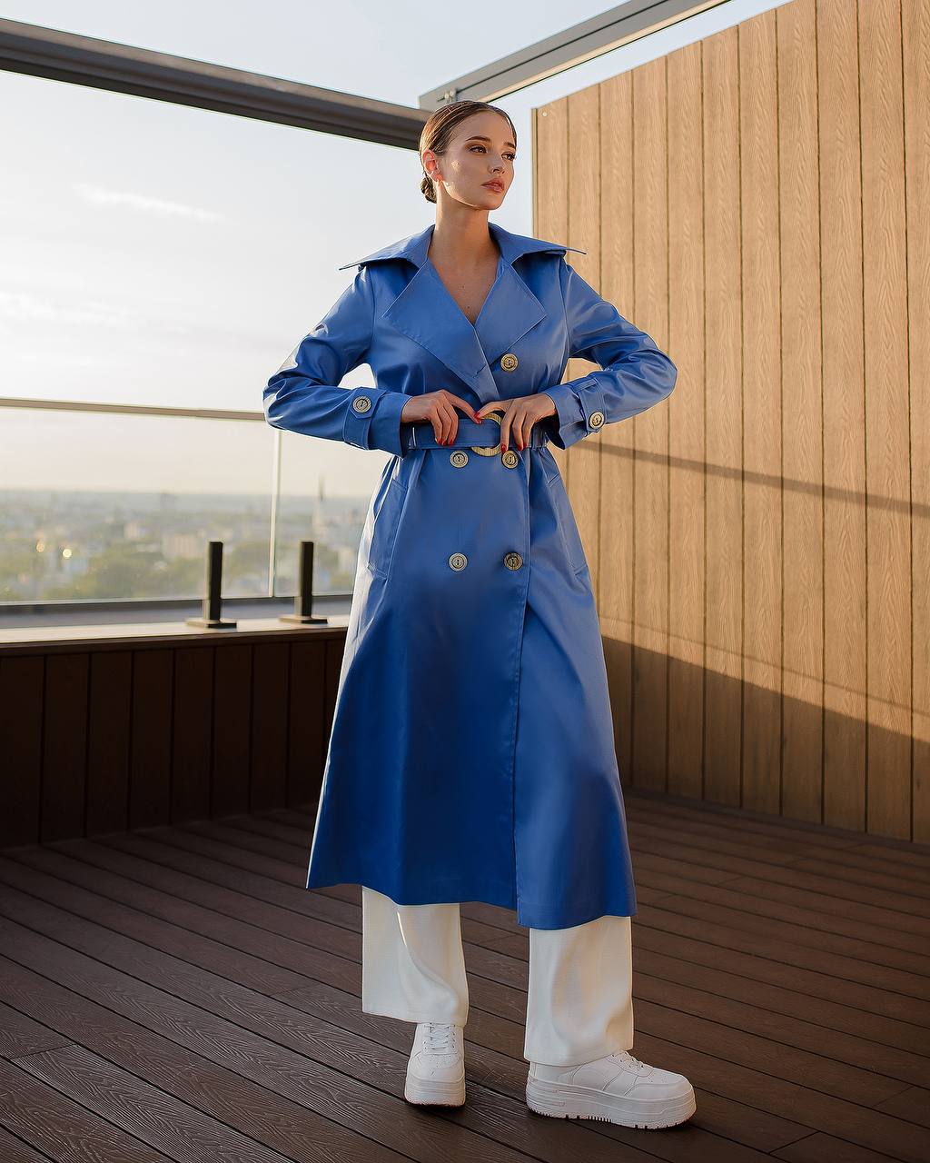 Person wearing a blue trench coat on a wooden deck with a cityscape view.