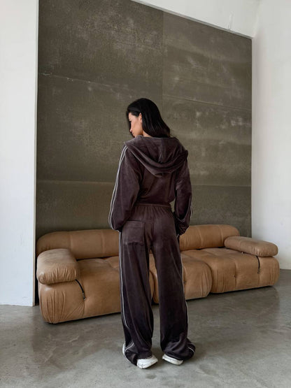 Person wearing a dark brown tracksuit standing in front of a brown sofa against a textured wall.