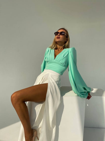 Woman in a turquoise top and white pants posing against a neutral background