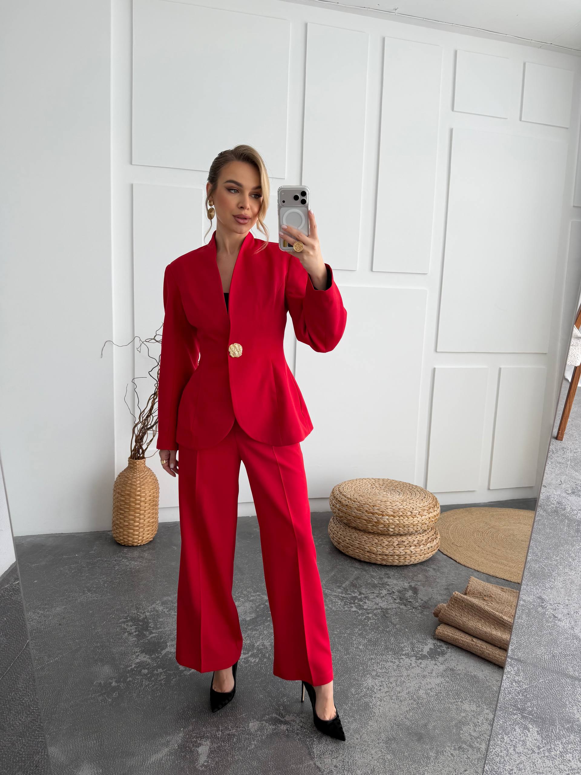 Person wearing a red suit taking a mirror selfie in a room with a white paneled wall and woven ottomans.