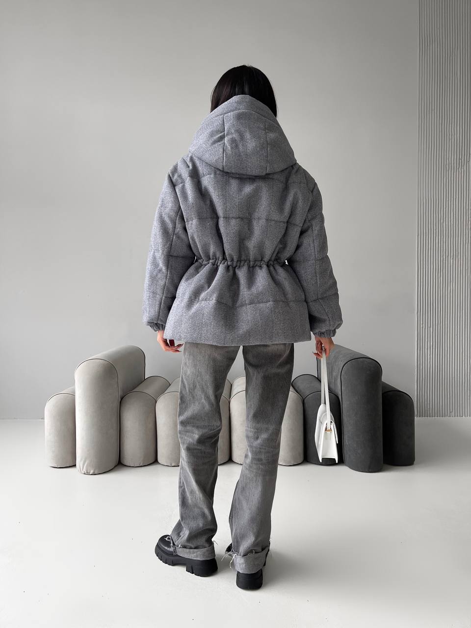Person wearing a gray hooded coat and pants standing in a minimalistic room with gray furniture.