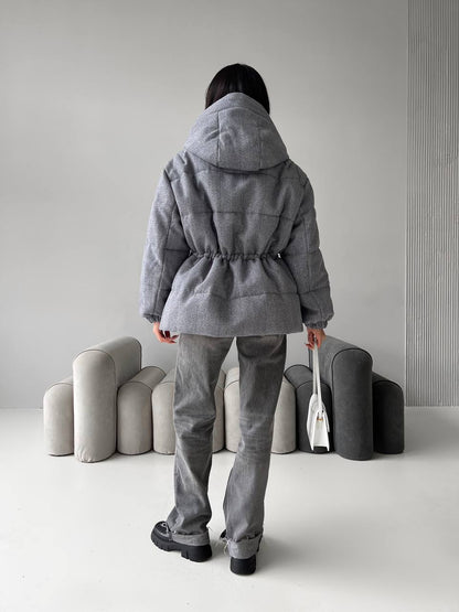 Person wearing a gray hooded coat and pants standing in a minimalistic room with gray furniture.