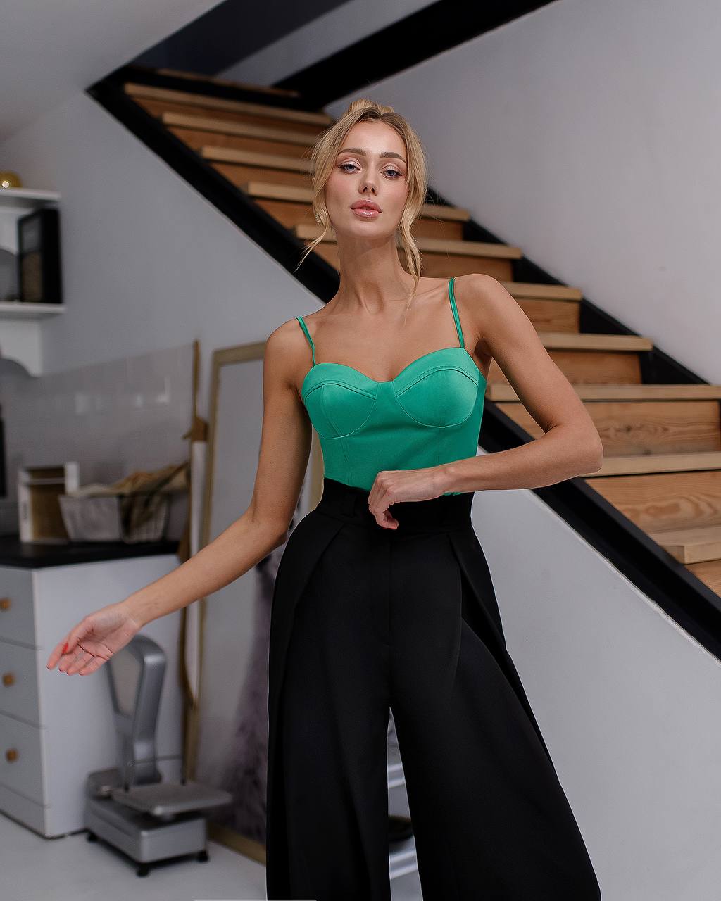 Woman in a green top and black pants standing in a room with wooden stairs.
