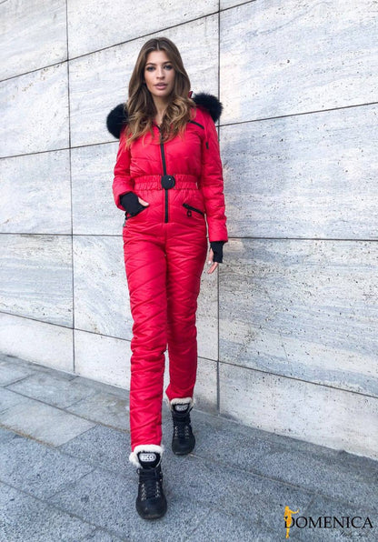 Woman wearing a red snowsuit with black fur trim against a gray concrete wall.