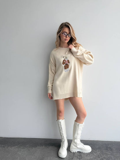 Woman wearing a beige sweater with a graphic design and white boots against a plain background