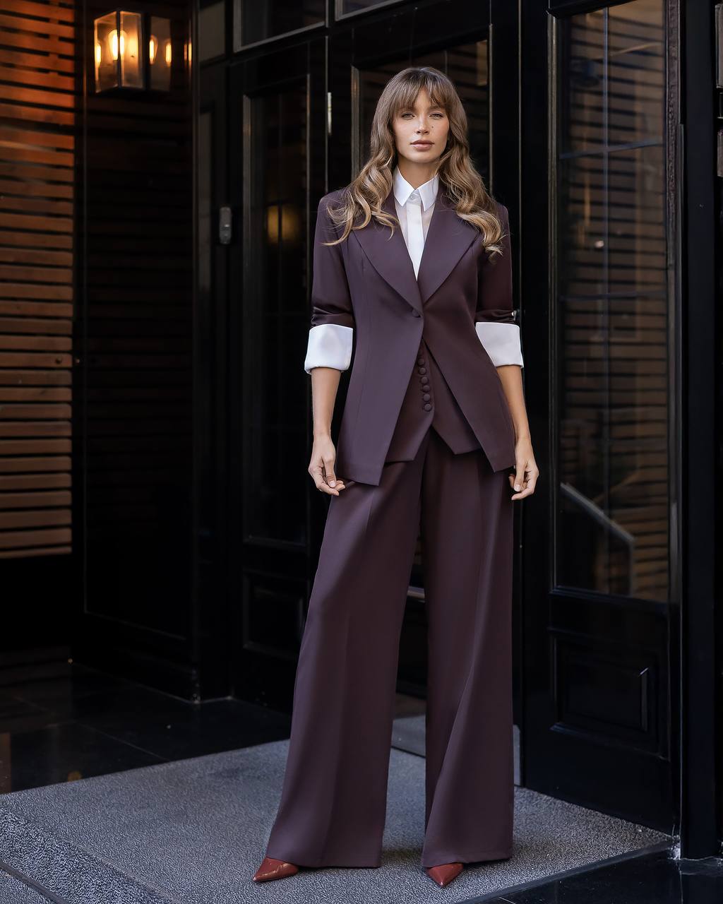Woman in a brown suit standing in front of a dark door.