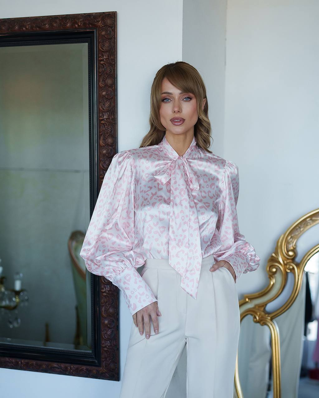 Woman wearing a pink floral blouse and white pants standing in a room with decorative mirrors.