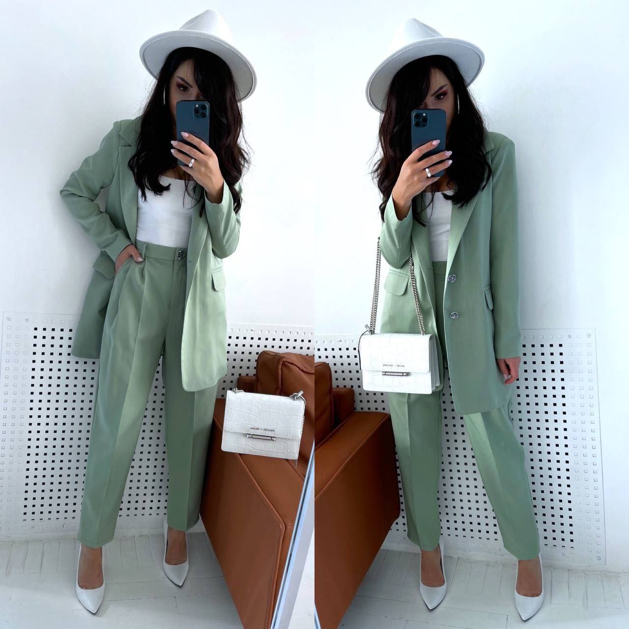 Woman in a green suit and white hat taking a mirror selfie in a minimalistic room.