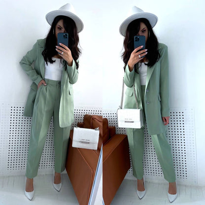 Woman in a green suit and white hat taking a mirror selfie in a minimalistic room.