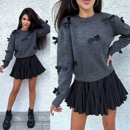 Woman wearing a gray sweater with black bows and a black skirt against a white wall.