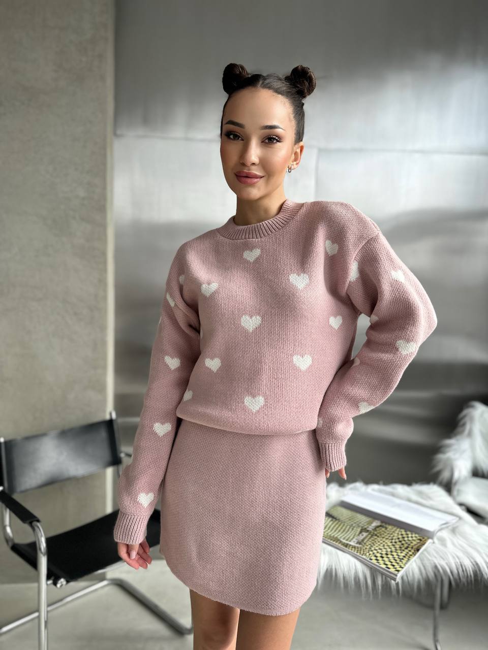 Woman wearing a pink sweater with heart patterns and matching skirt in a room.