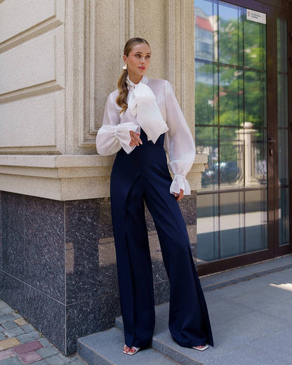 Woman in a white blouse and navy pants standing against a building.