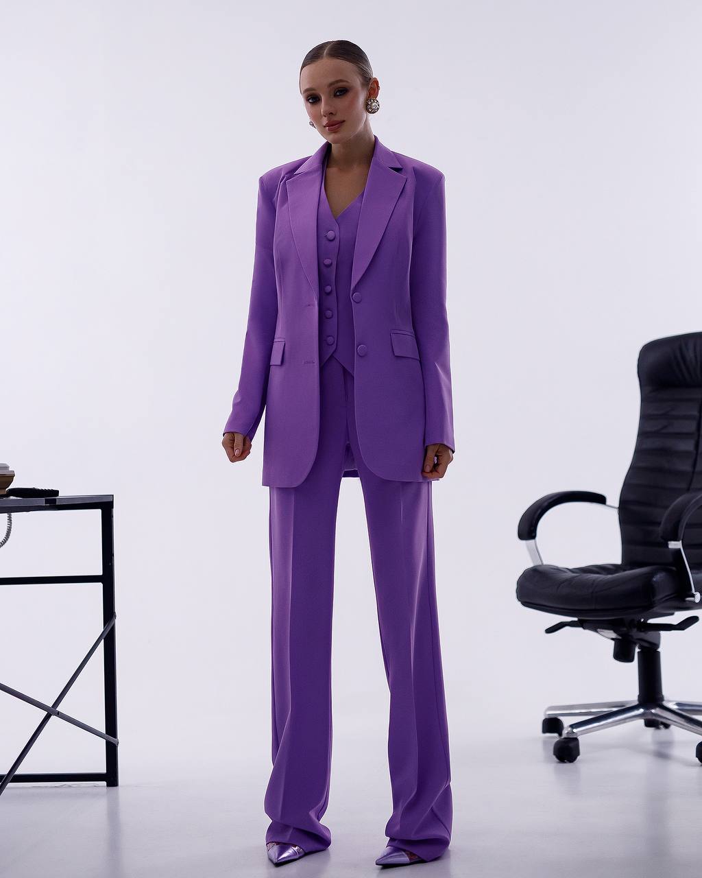 Woman wearing a purple suit standing in an office setting with a chair and desk.