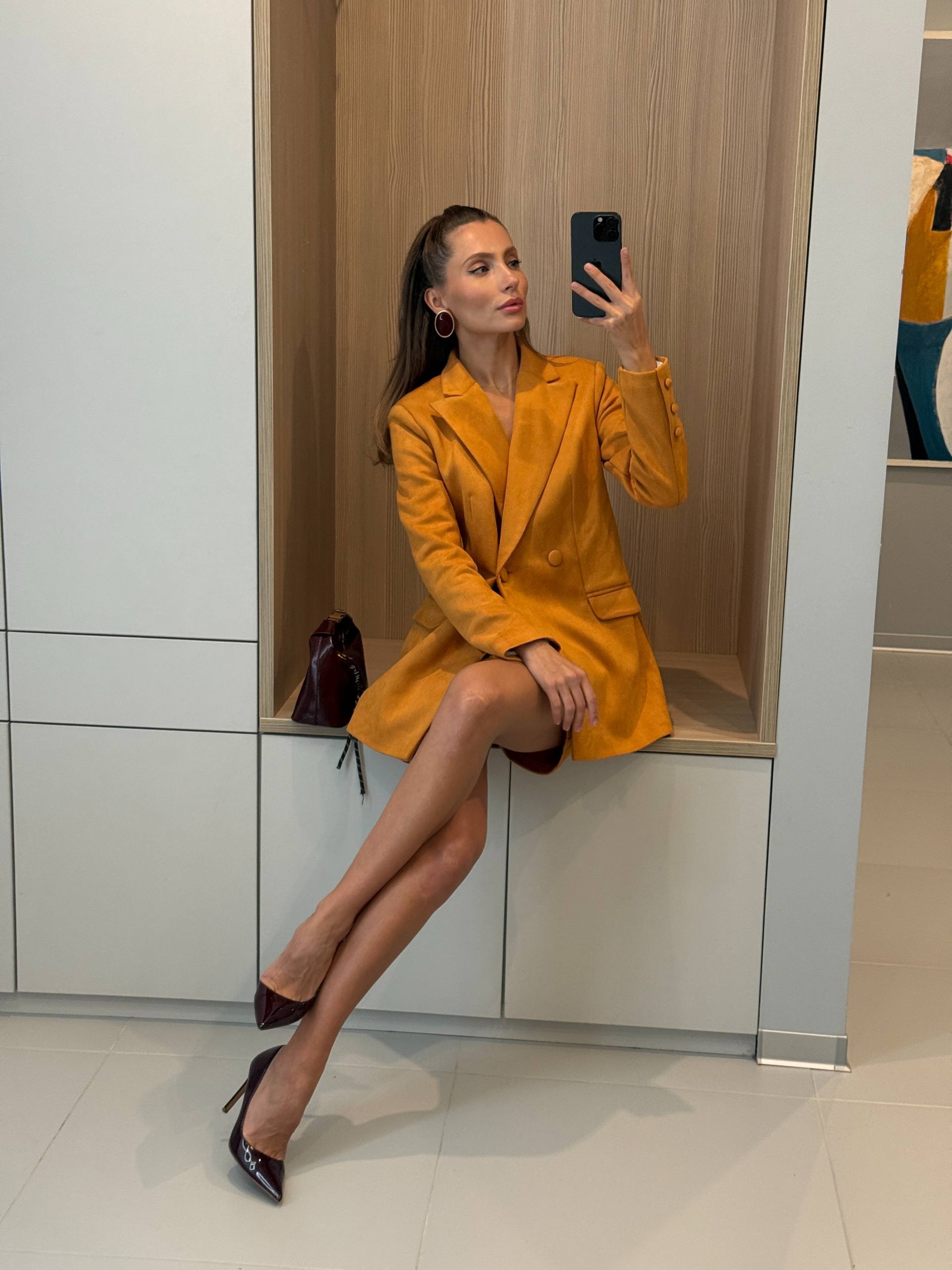 A woman takes a mirror selfie in a clothing store while wearing a mustard yellow blazer dress, a dark red mini dress, and dark brown pointed-toe heels.