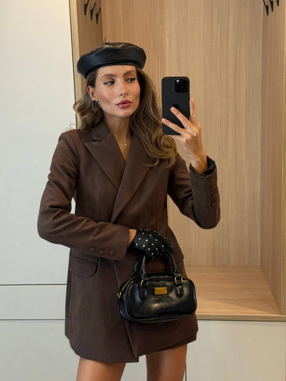 A woman takes a mirror selfie wearing a brown double-breasted blazer and matching mini skirt, a black beret, black studded gloves, and holding a black handbag with gold hardware.