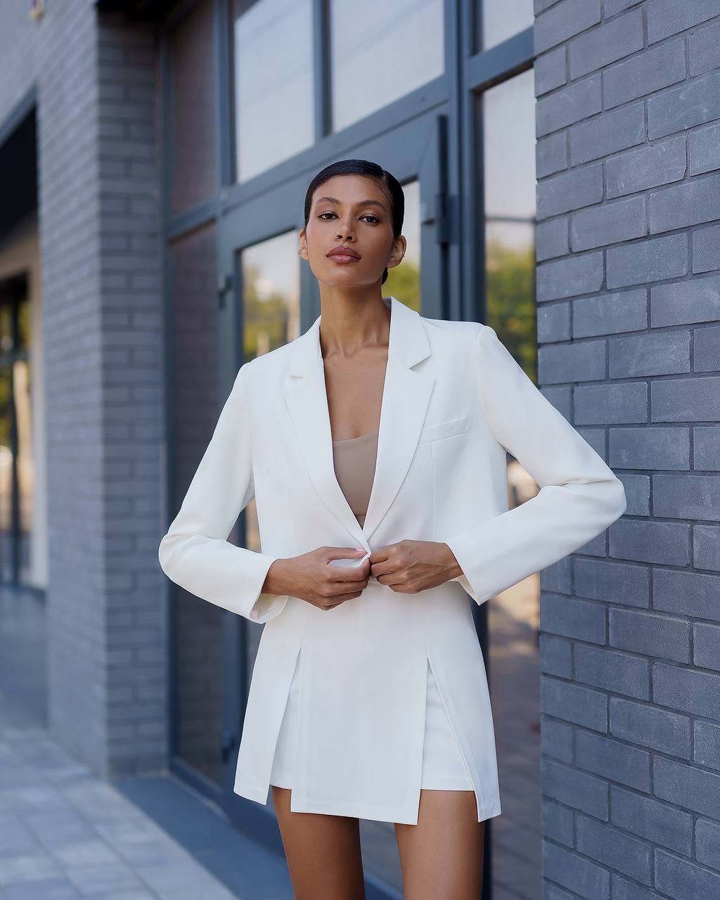 Woman wearing a white blazer over a beige top and skirt standing against a brick wall.