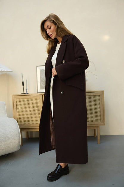 Woman wearing a long dark brown coat in a room with a light-colored wall and furniture.