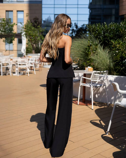 A woman stands on an outdoor rooftop patio wearing a black sleeveless top and wide-leg pants, photographed from behind in bright sunlight.