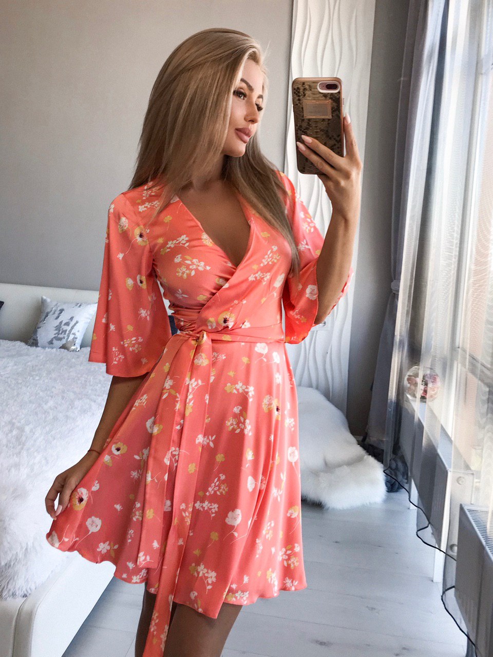 Woman taking a mirror selfie wearing a coral floral dress in a bedroom.