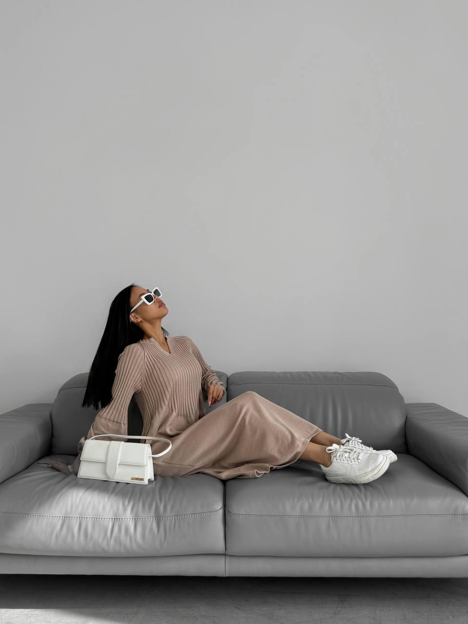 Woman sitting on a gray sofa wearing sunglasses and holding a white handbag against a plain background
